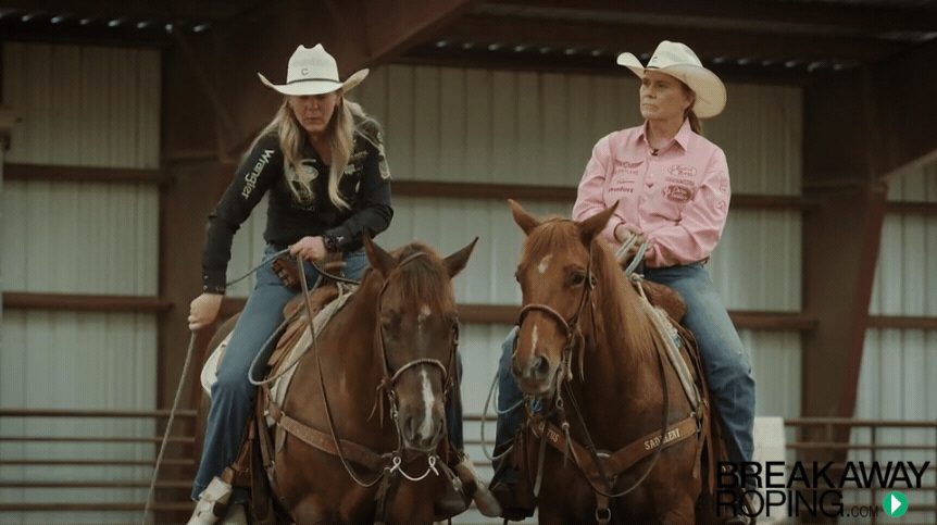 Two women in cowboy hats on saddled horses in an indoor arena with "Breakaway Roping" text visible