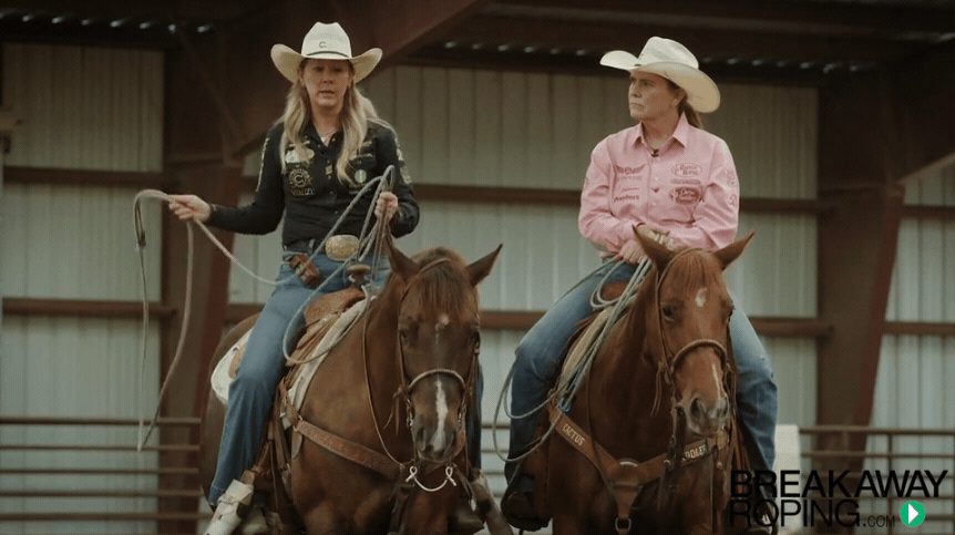 Two female roping competitors on horseback in an indoor arena, one holding a lasso, both wearing cowboy hats and western attire