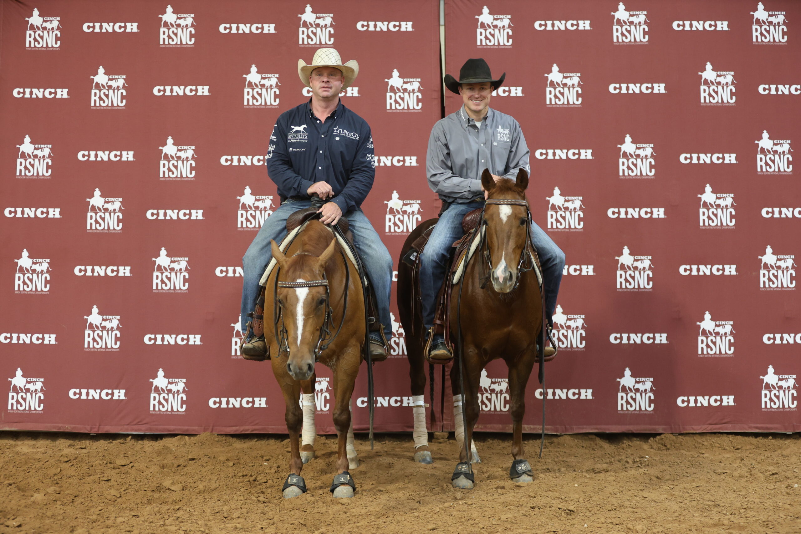 Two mounted riders in cowboy hats pose side by side on sorrel horses in front of a Cinch RSNC backdrop