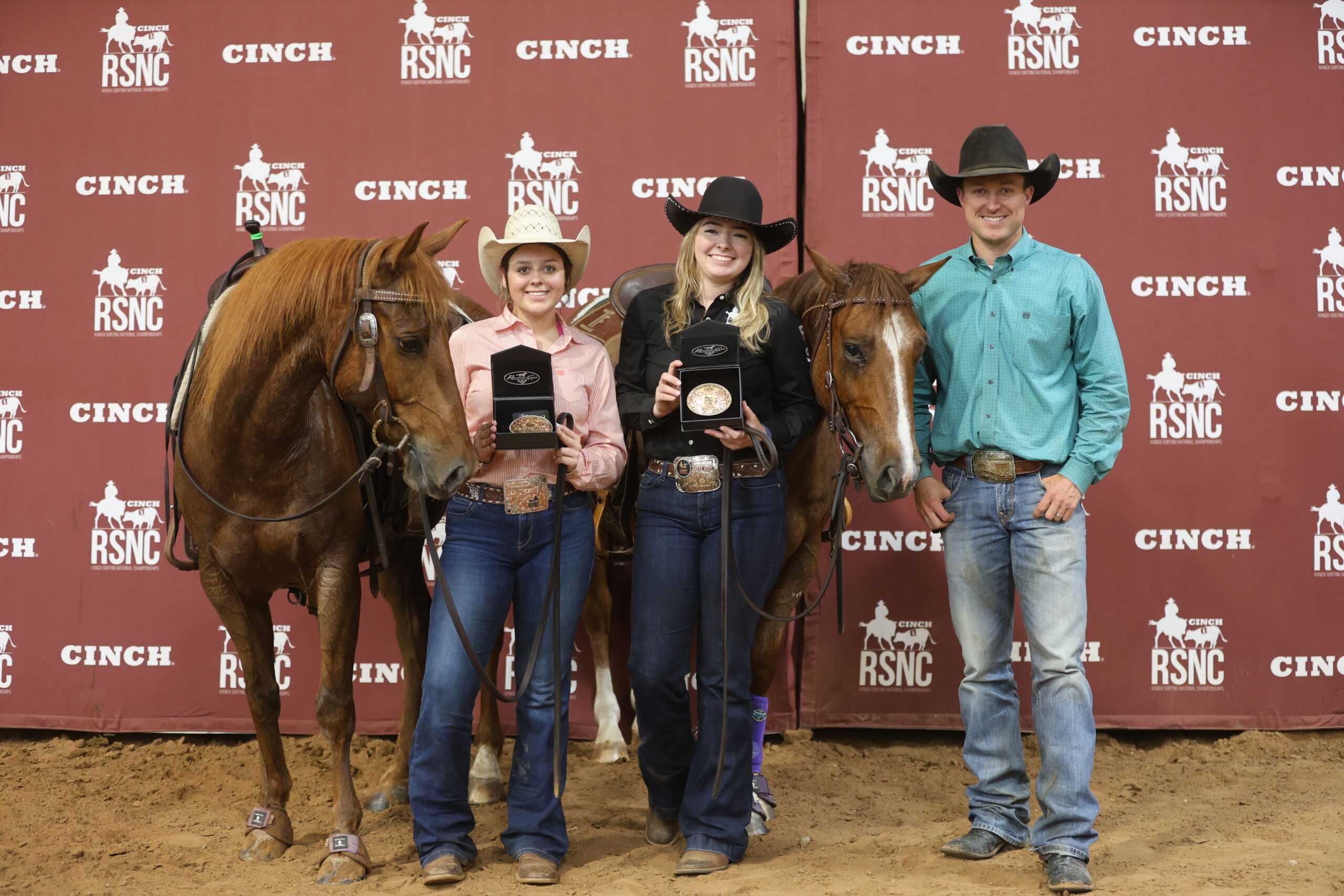 Two young women in cowboy hats hold championship buckles beside their sorrel horses as a man in a turquoise shirt stands at right in front of a Cinch RSNC backdrop