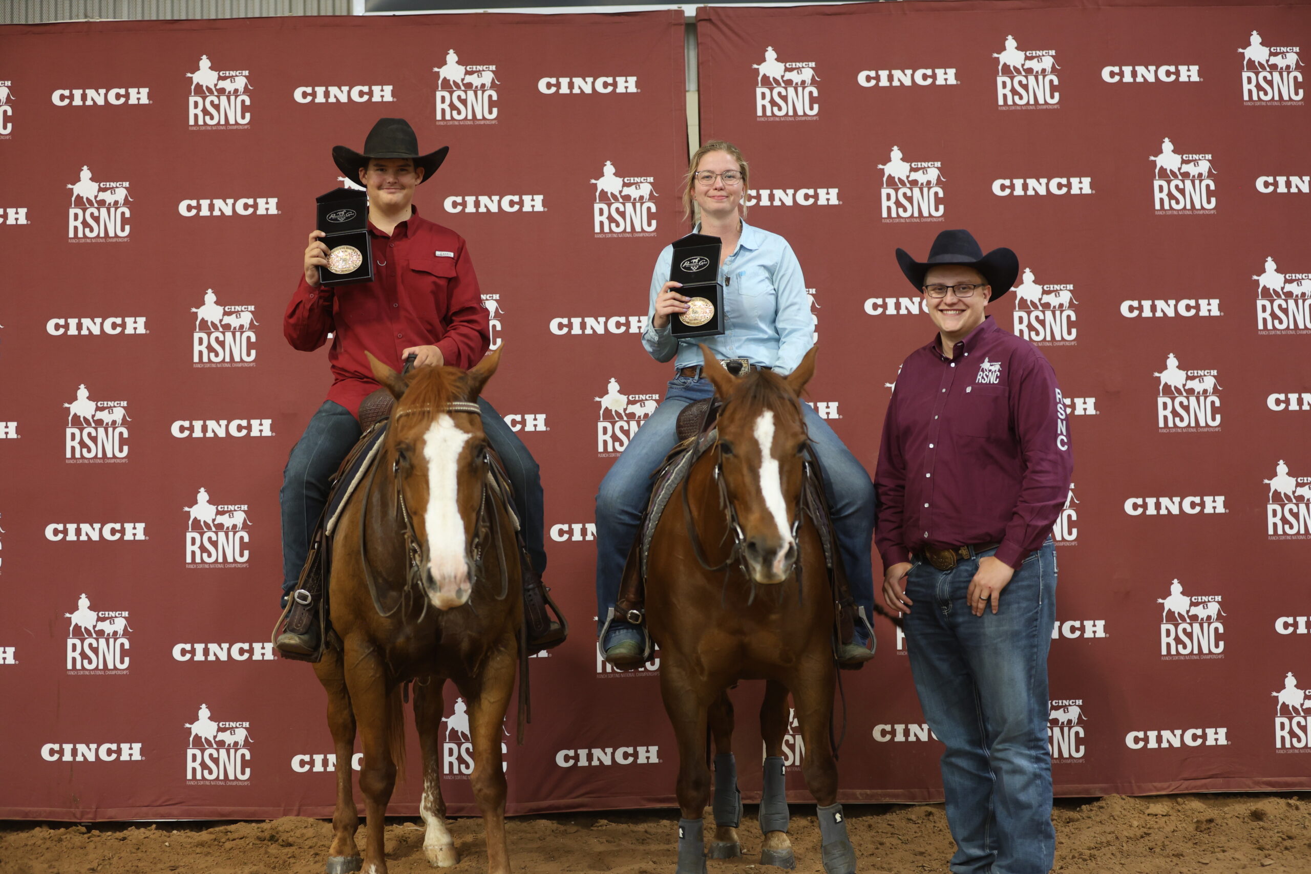 Two mounted ranch sorting competitors hold championship buckles beside a man in a maroon RSNC shirt at a Cinch RSNC backdrop