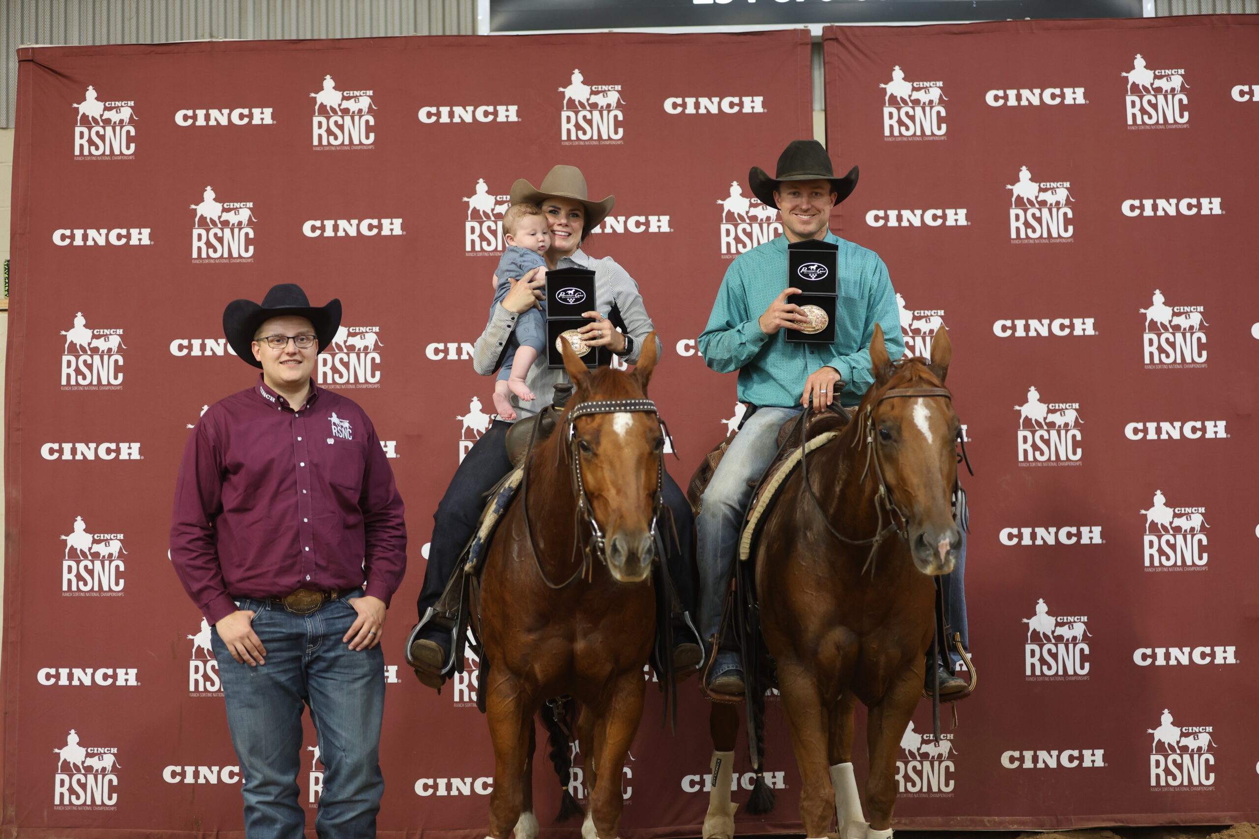 Two mounted riders, one holding a baby, display trophy buckles beside a man on foot in front of a Cinch RSNC backdrop