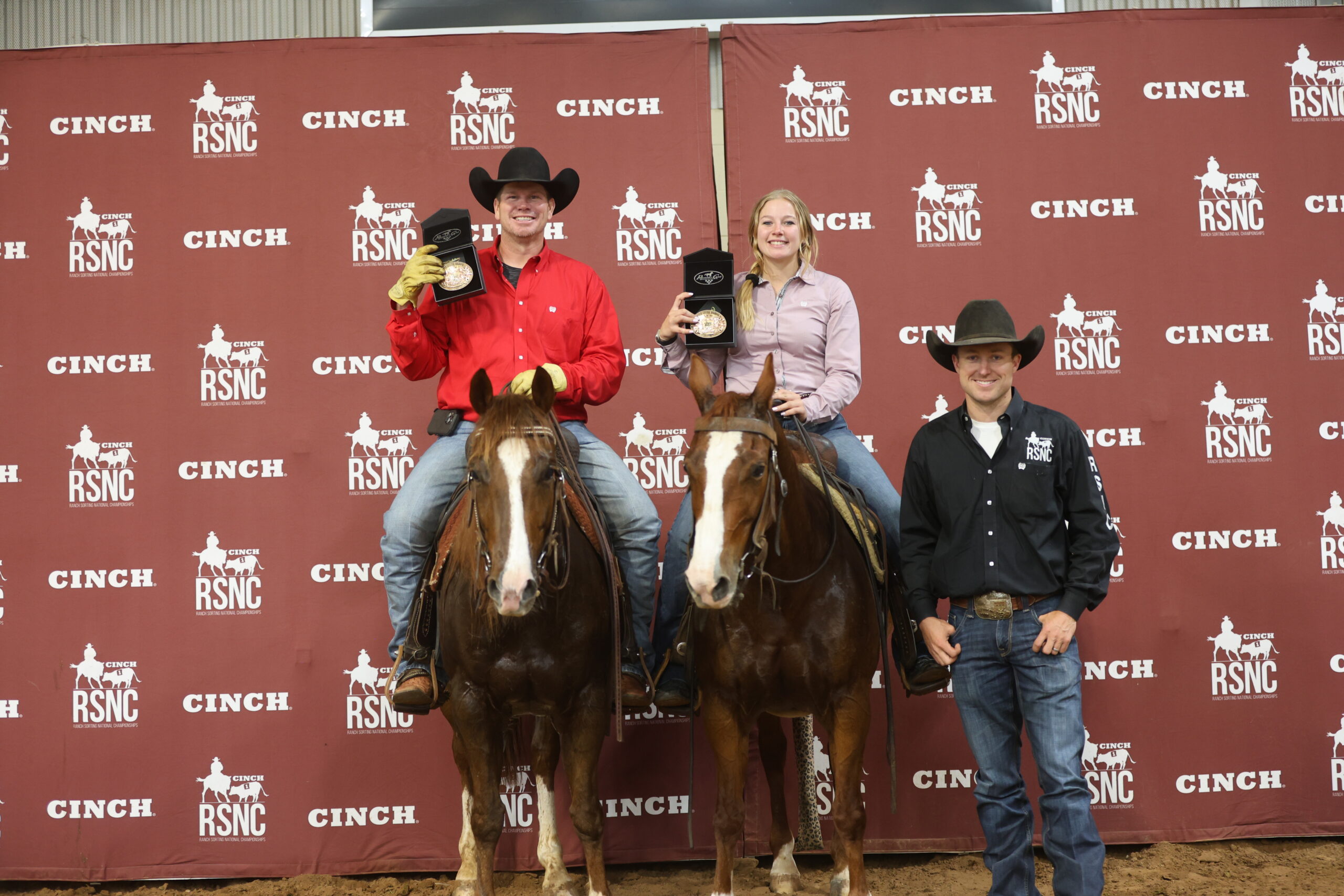 Two mounted ranch sorting winners hold up buckle boxes beside a standing official in front of a Cinch RSNC backdrop