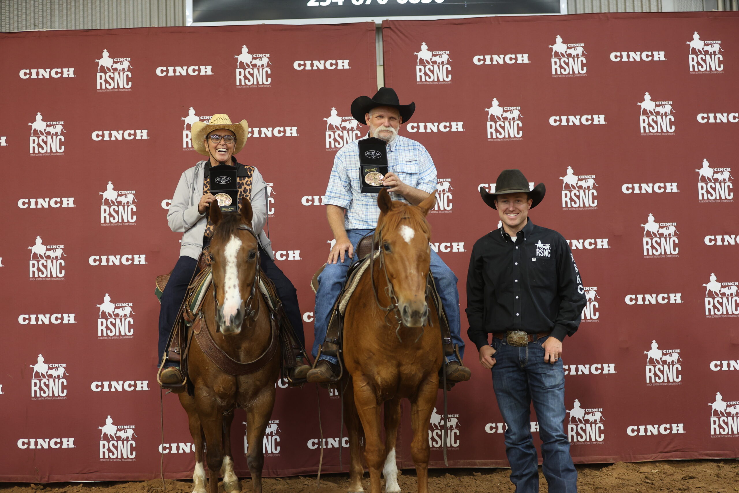 Two mounted riders hold up championship buckles beside a standing man in an RSNC shirt in front of a Cinch RSNC backdrop