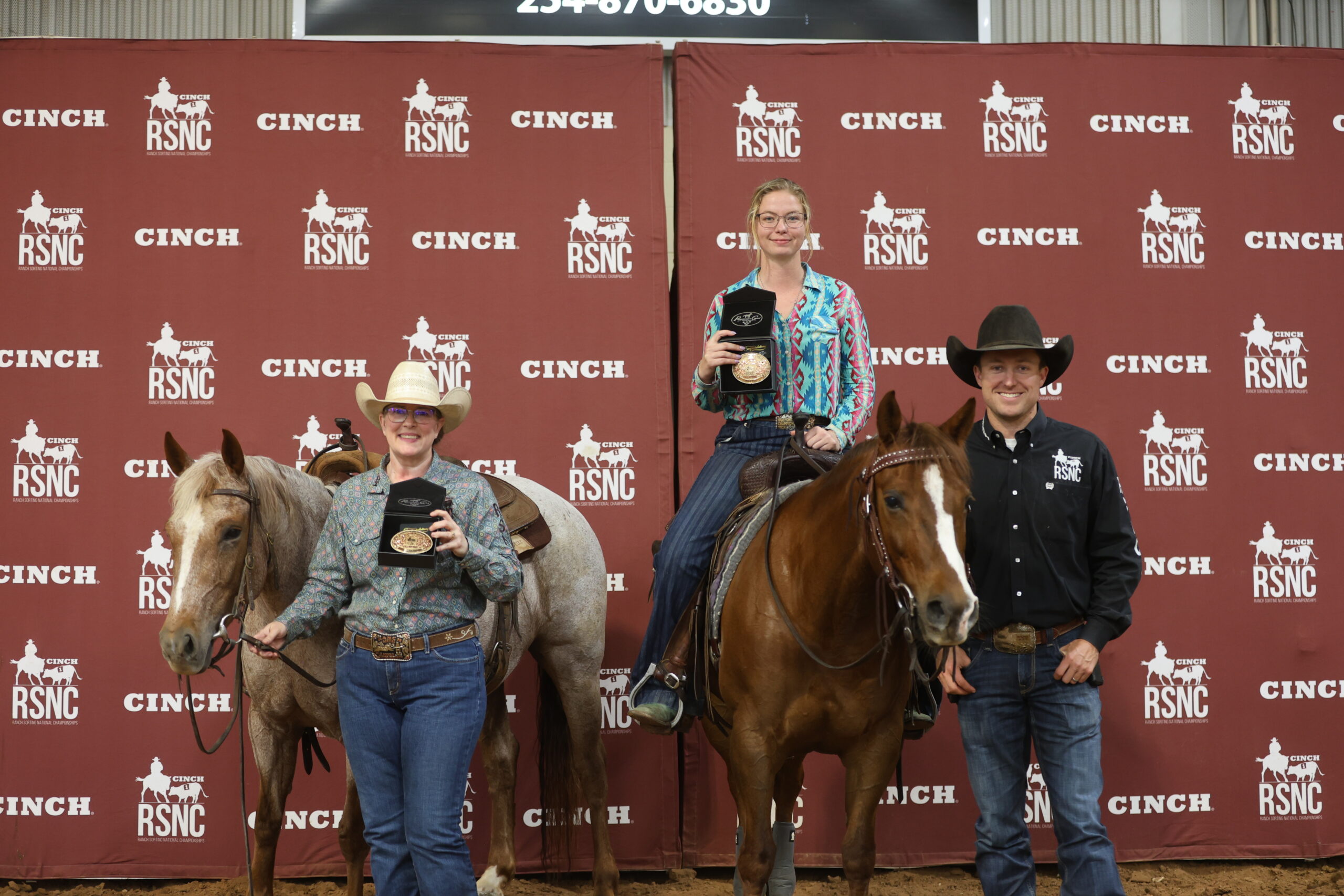 Two women hold championship buckles, one standing beside a roan horse and one mounted on a sorrel, next to a man in a cowboy hat at a Cinch RSNC backdrop