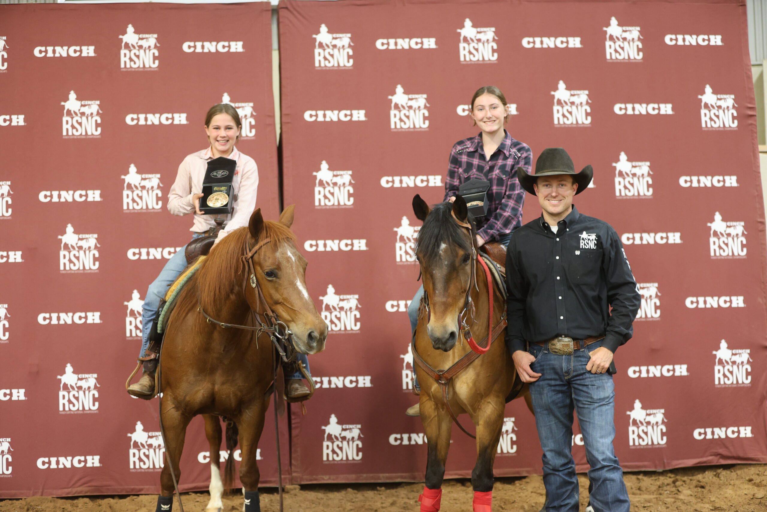 Two young riders hold trophy buckles horseback beside a man in a black cowboy hat in front of a Cinch RSNC backdrop