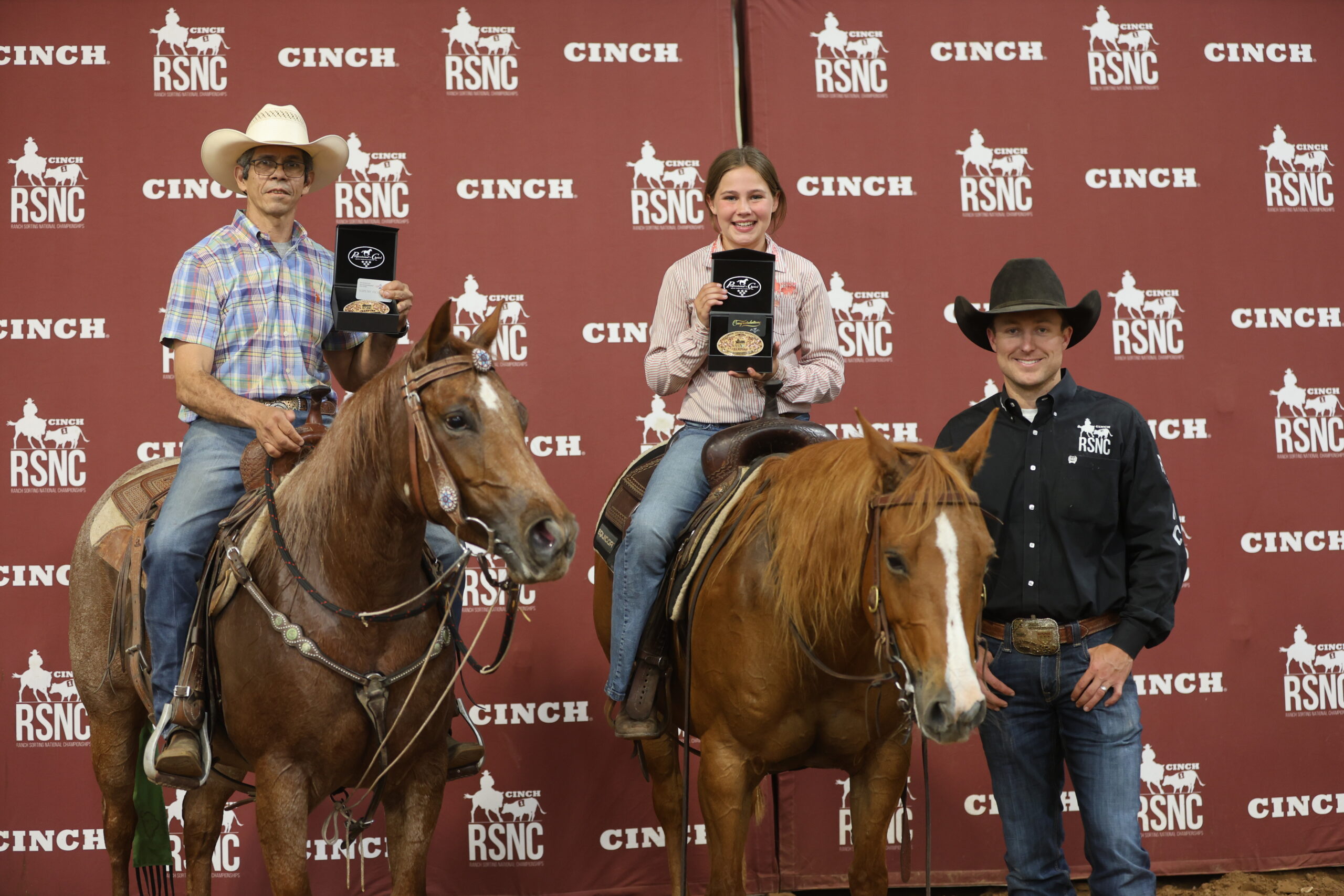 Two mounted riders hold championship buckles in presentation boxes beside a standing man in front of a Cinch RSNC backdrop