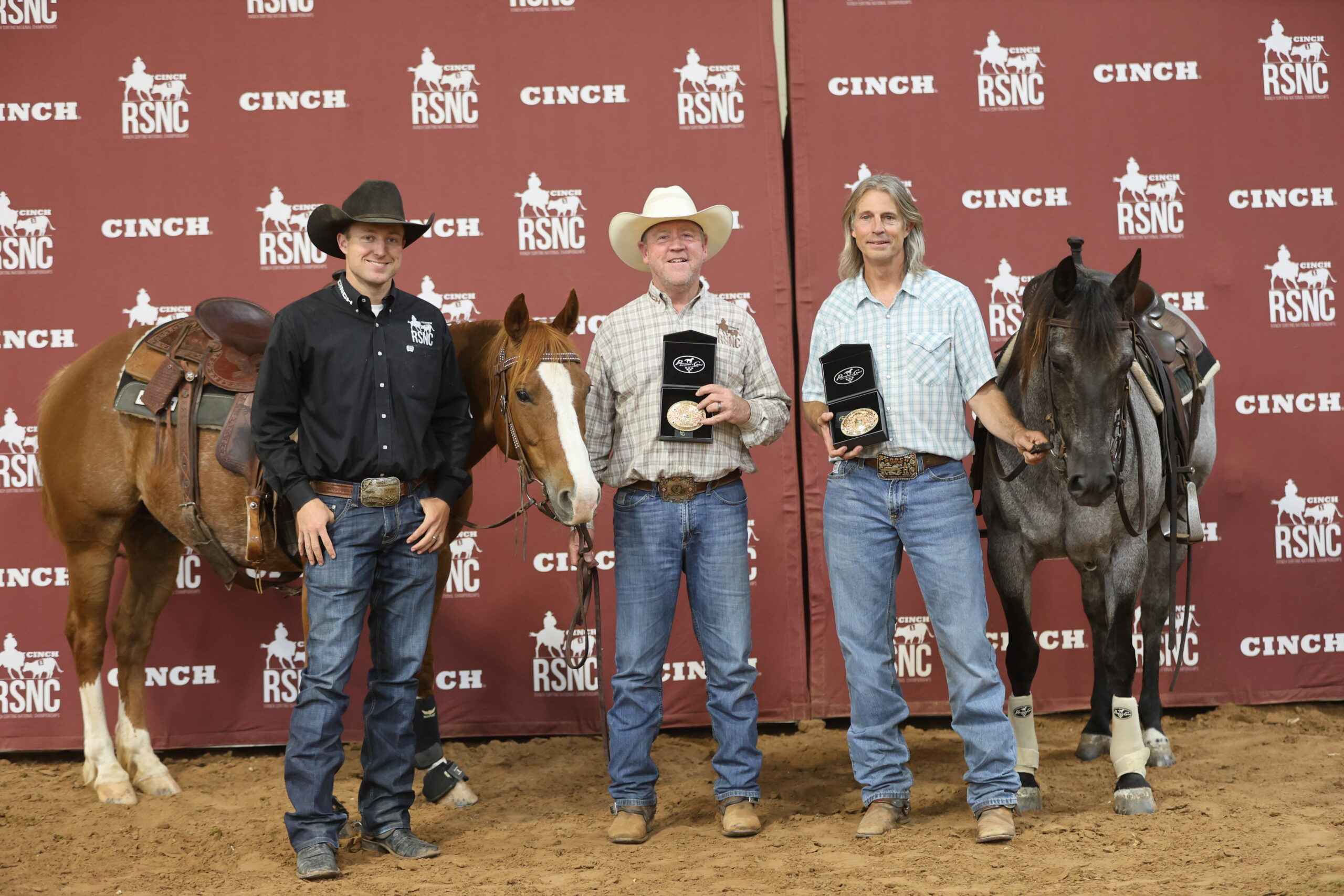 Two riders hold trophy buckles beside their saddled sorrel and blue roan horses in front of a Cinch RSNC backdrop