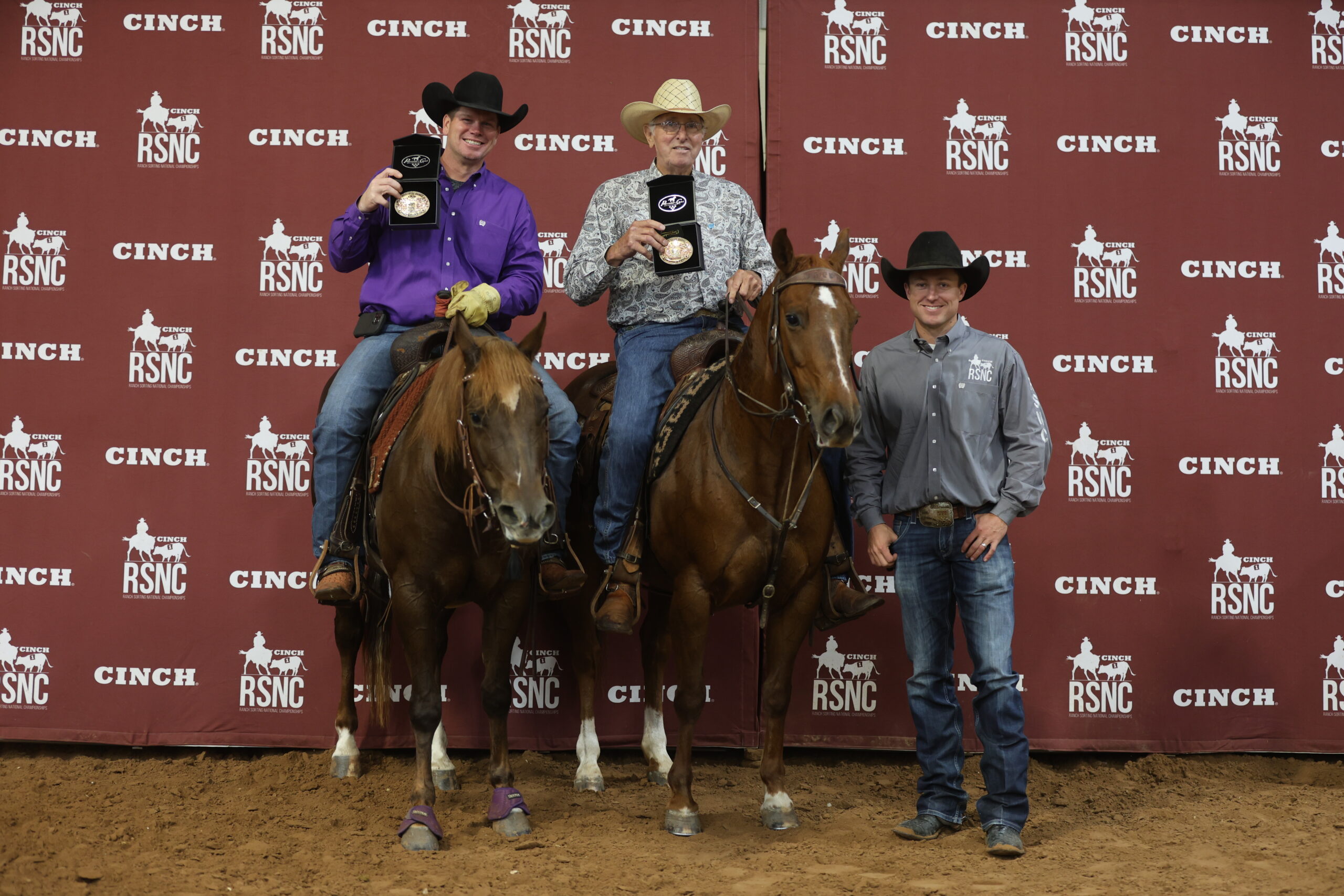Two mounted ranch sorting partners hold up championship buckles beside a standing official in front of a Cinch RSNC backdrop