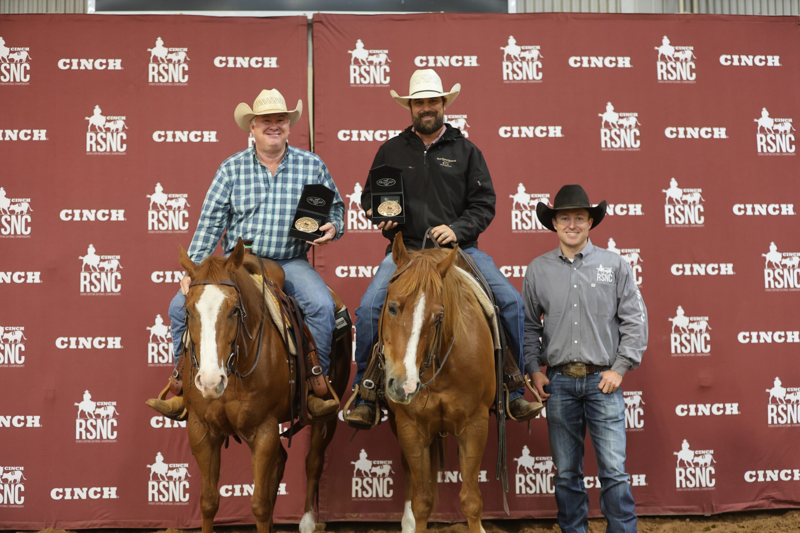 Two mounted riders hold championship buckles in presentation cases beside a standing man in front of a Cinch RSNC backdrop