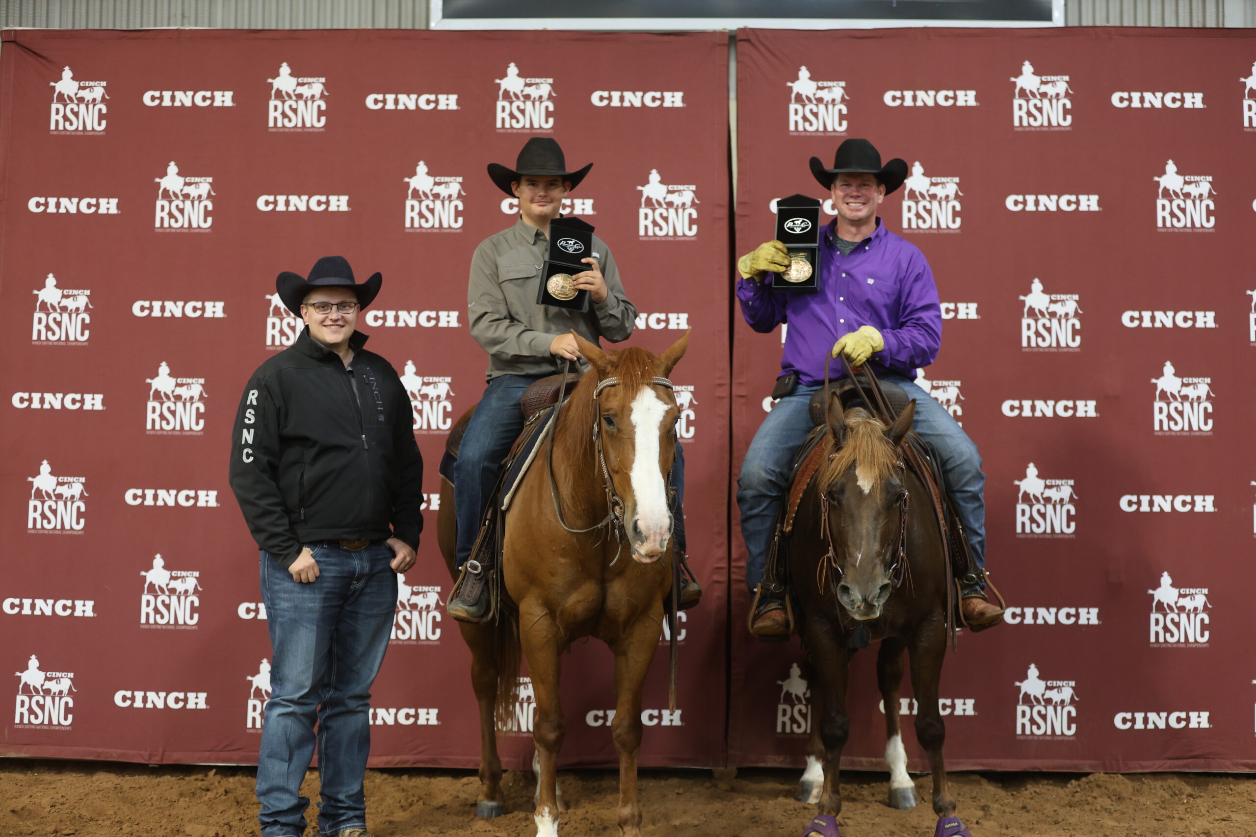 Two mounted riders hold up championship buckles beside a standing man in an RSNC jacket in front of a Cinch RSNC backdrop