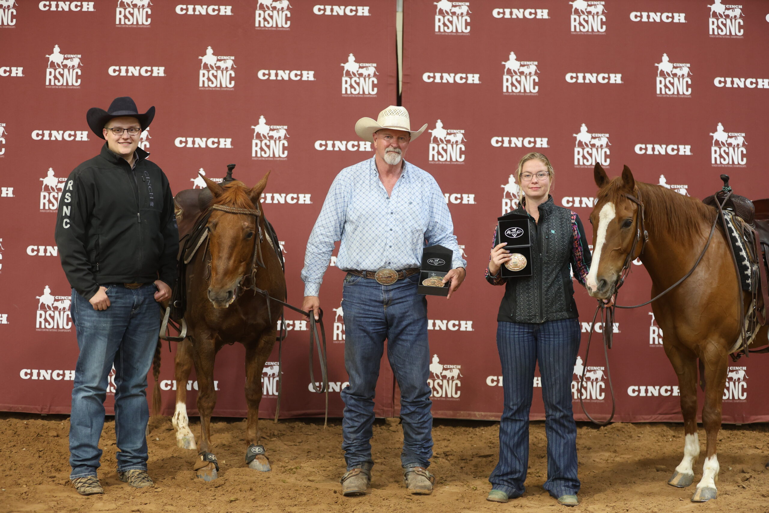 Two men and a woman hold trophy buckles beside two sorrel horses in front of a Cinch RSNC backdrop