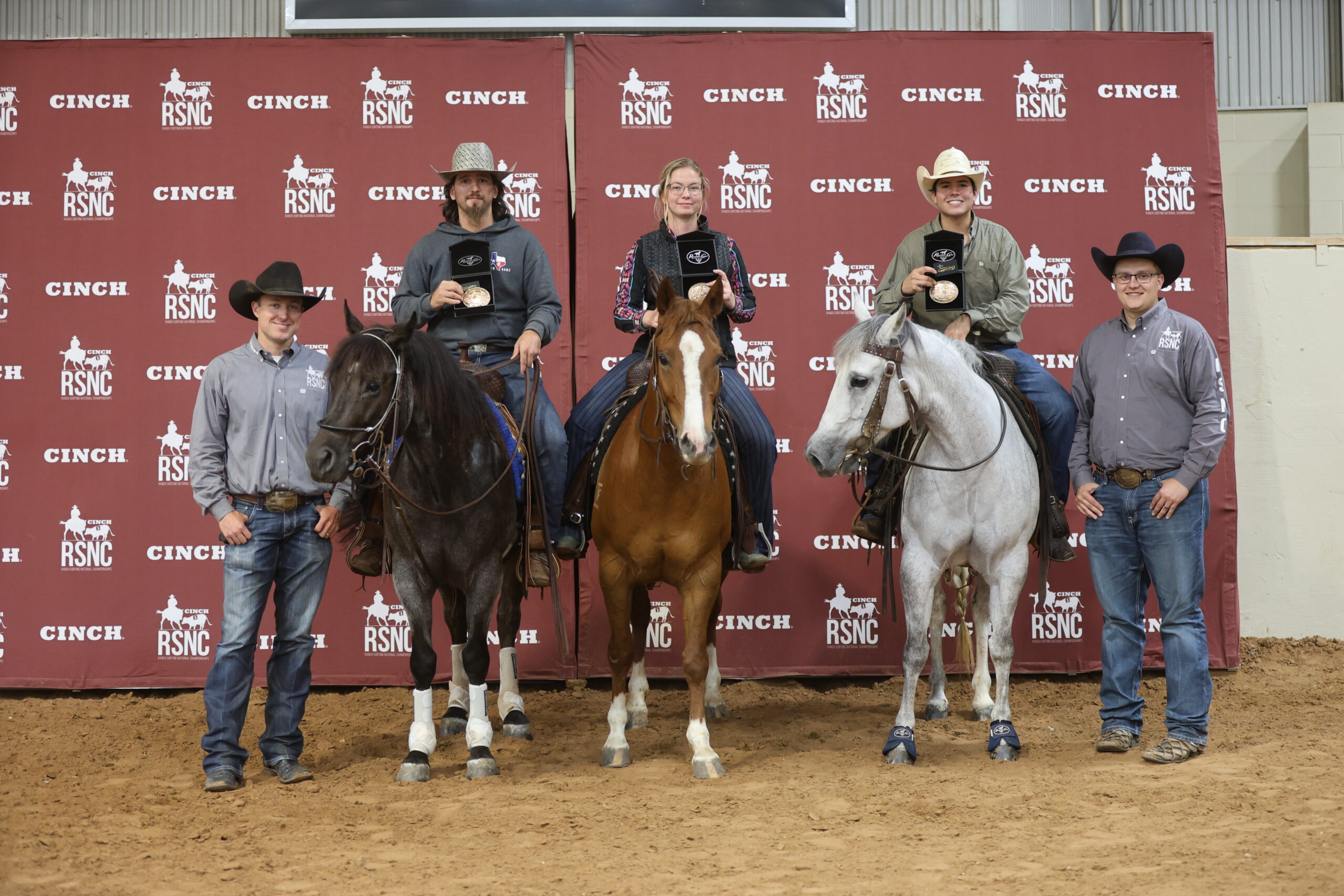 Three mounted riders hold buckle presentation boxes beside two men on foot in front of a Cinch RSNC backdrop