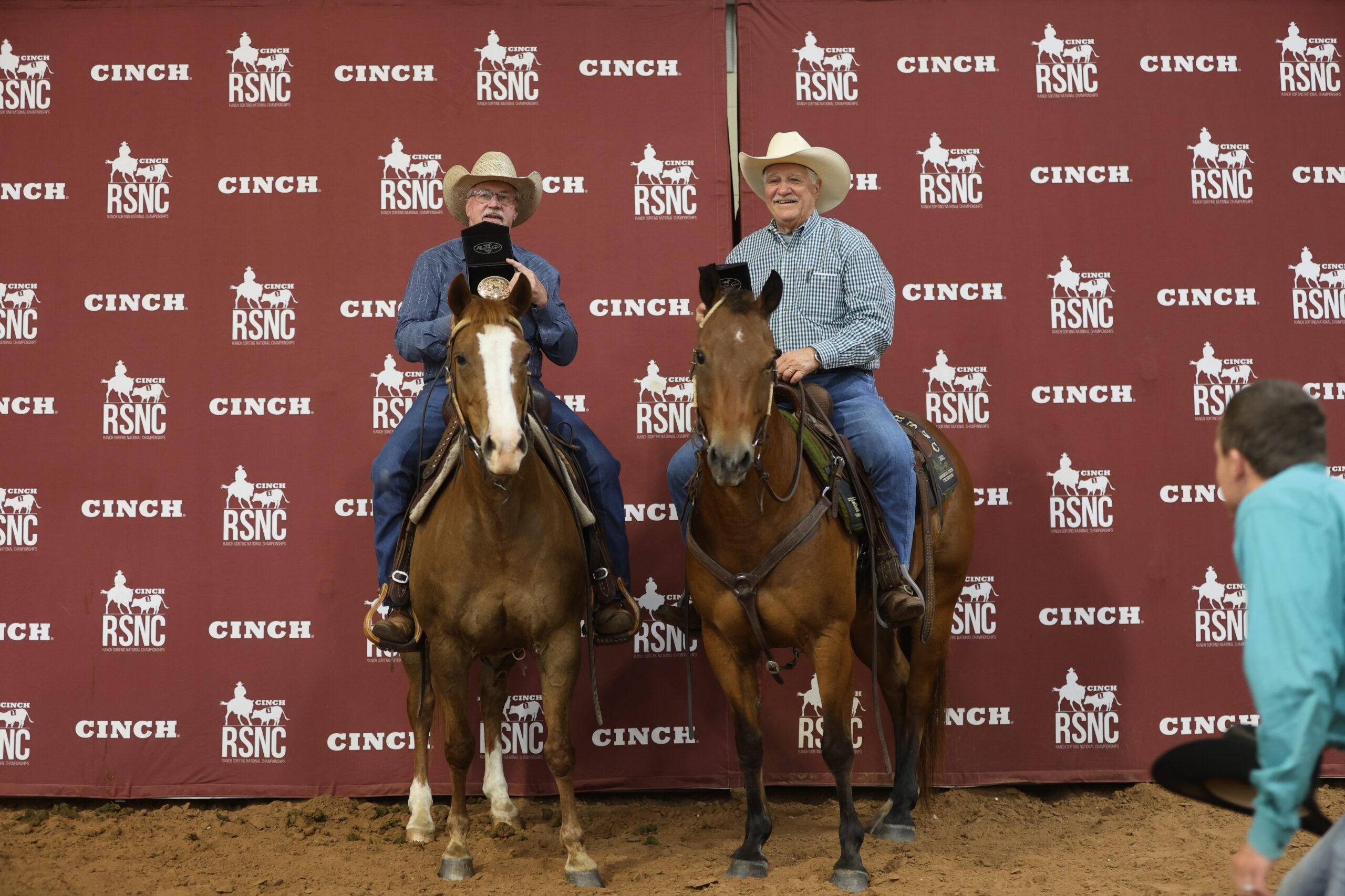Two smiling cowboys on horseback hold up championship buckle boxes in front of a Cinch RSNC backdrop