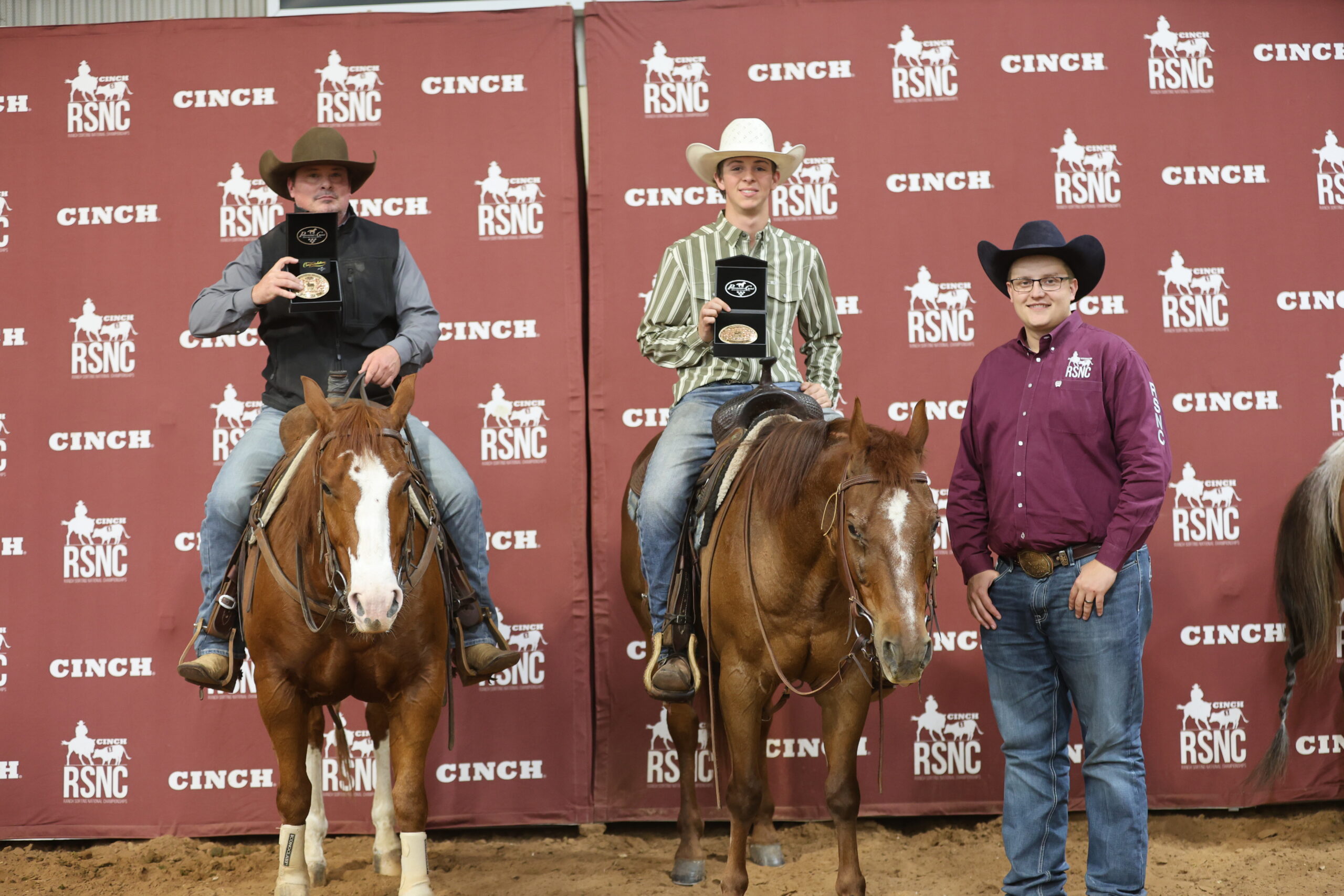 Two mounted riders hold championship buckles in presentation boxes beside a man in a maroon RSNC shirt at a Cinch RSNC backdrop