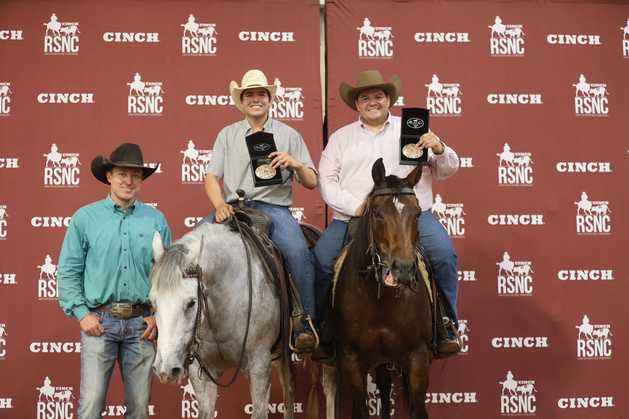 Two mounted cowboys hold up trophy buckles in presentation boxes beside a man standing at a gray horse's head, in front of a Cinch RSNC backdrop