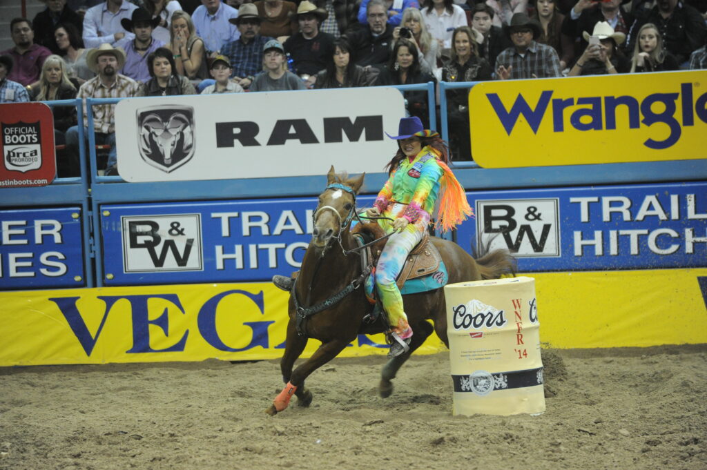 Barrel racer on horseback rounds a yellow barrel during competition, with arena sponsors and crowd visible in background