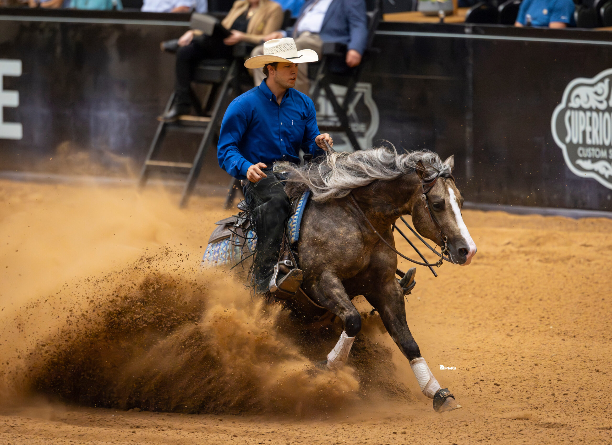 Reiner in a blue shirt and cowboy hat slides a dapple gray horse to a stop, kicking up a wall of arena dirt